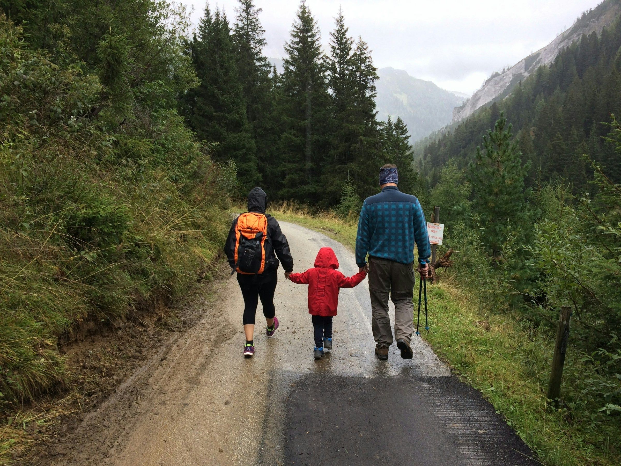 A family of three walks a mountain trail together, the child in a red jacket between two adults